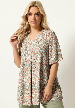 Blond kvinde med løse bølger bærer en blomsterkortærmet tunika med pink og orange blomster, stående mod en ensfarvet baggrund.