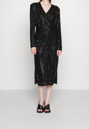 Vrouw die een zwarte midi-jurk met lange mouwen en pailletten draagt, met een geplooide voorkant en zwarte open-toe muiltjes met hak, staand tegen een effen achtergrond.