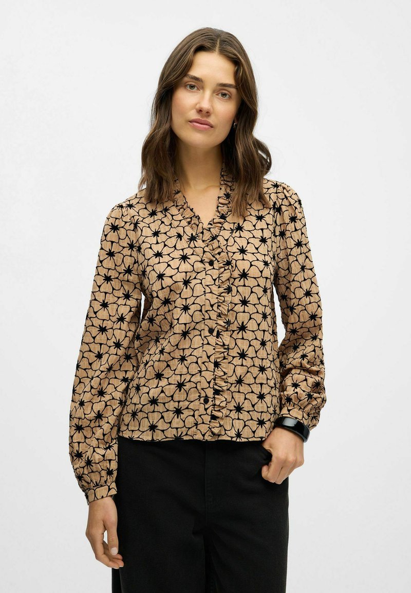 Blusa marrón con patrón y diseño floral negro, con cuello rizado y mangas largas. La tela parece ligera y tiene un acabado texturizado.