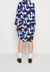 Dlouhá košilová šaty s výrazným abstraktním vzorem v černé, modré a krémové barvě. Vyrobeno z lehkého materiálu, s volným střihem a dlouhými rukávy.