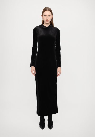 Čern dress s kapucí z sametu, dlouhými rukávy, přiléhavým siluetu a dlouhým lemem až na zem. Modelka má na sobě špičaté černé boty.