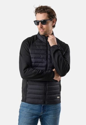 Man with light brown hair wearing black sunglasses, black quilted jacket, and blue jeans, posing with one hand near his neck.
