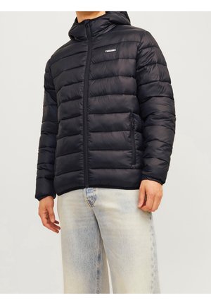 Homme portant une veste matelassée noire à capuche avec des poches zippées et un jean délavé clair sur fond blanc.