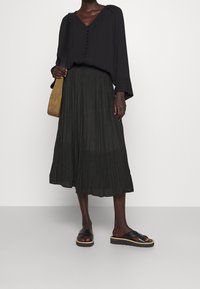 Schwarze Bluse mit langen Ärmeln, V-Ausschnitt und Knopfdetails, kombiniert mit einem plissierten schwarzen Rock und schwarzen Sandalen mit dicker Sohle.