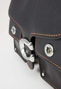 Gros plan sur un sac en cuir noir avec des coutures marron et des détails en métal argenté sur le fermoir et les boutons, sur fond blanc.
