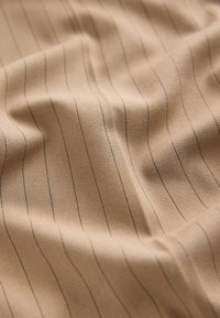 Beige Stoff mit glatter Textur weist dünne, vertikale schwarze Streifen auf. Das Material wirkt leicht und fällt sanft.
