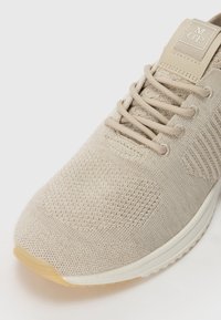 Beige treningssko med et strukturert strikket overdel, forsterket tåhette og flate lisser. Har en lett gummisåle og logo på tungen.