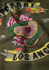 Tessuto verde oliva con motivo di serpente e fiori con ornamenti colorati. Il testo include "LOS ANGELES" su un banner.