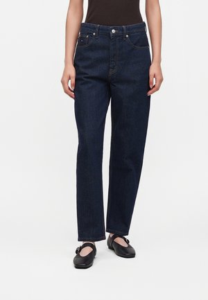 Person iført mørkeblå jeans med lige ben, sort top og sorte flade sko med spænder, stående mod en ensartet hvid baggrund.