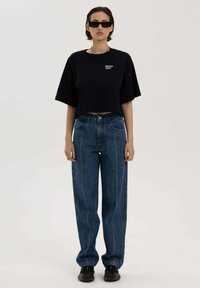 Zwarte cropped t-shirt met logo, blauwe high-waisted jeans met zichtbare stiksels, en zwarte vierkante zonnebril gedragen met chunky zwarte schoenen.
