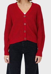 Cardigan rouge en tricot avec un col en V, cinq boutons, poignets côtelés et un ourlet légèrement court. Porté sur un jean noir taille haute.