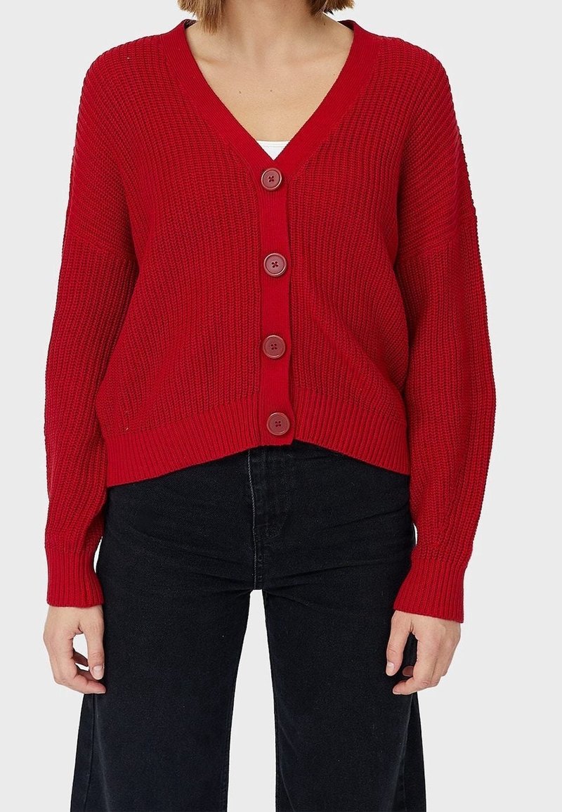 Cardigan rouge en tricot avec un col en V, cinq boutons, poignets côtelés et un ourlet légèrement court. Porté sur un jean noir taille haute.