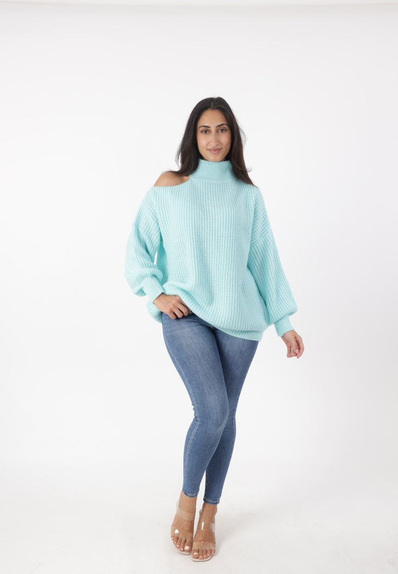 Maglione azzurro chiaro a maglia con spalle scoperte, collo alto e maniche ampie, abbinato a jeans blu aderenti e sandali con tacco a blocco trasparente.