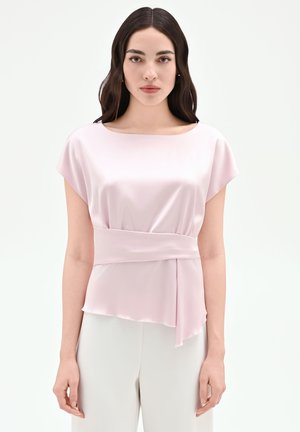 Donna con capelli lunghi e scuri che indossa una camicetta di raso rosa chiaro con dettaglio di cintura larga e pantaloni bianchi su uno sfondo uniforme.