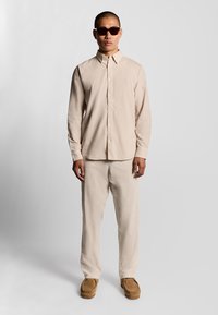 Mand iført beige knap-op skjorte, matchende bukser, brune mokkasin sko og mørke solbriller, står mod en enkel lys baggrund.