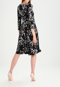 Robe noire à fleurs avec des manches longues, dotée d'une taille cintrée et d'une jupe évasée. Agrémentée de grands motifs de lys blancs. Portée avec des talons nus.