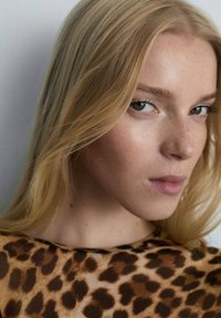 Frau mit langen, glatten blonden Haaren, die ein Oberteil mit Leopardenmuster trägt. Das Oberteil hat ein weiches, strukturiertes Muster in Brauntönen und Schwarz.