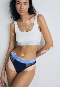 White crop top with gray "HUGO" brand logo, paired with navy briefs featuring a blue polka dot waistband and "HUGO" branding.