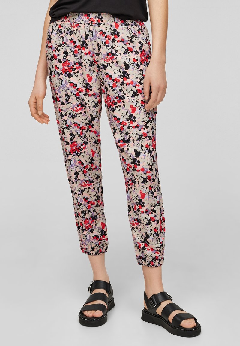 Pantalon à imprimé floral avec une base rose clair, comportant des fleurs rouges, noires et violettes ; taille élastique ; jambe fuselée ; porté avec des sandales noires.