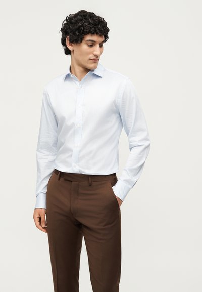 Άνδρας που φορά ανοιχτό μπλε ριγέ πουκάμισο με κουμπιά και καφέ παντελόνι, στέκεται με ένα χέρι στην τσέπη, κοιτάζοντας προς τα κάτω πλάγια.