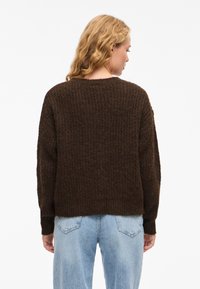 Maglione marrone lavorato a maglia con motivo testurizzato, scollo rotondo e vestibilità ampia. Polsini delle maniche e orlo leggermente a coste. Indossato con jeans azzurri.