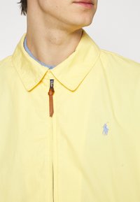 Gelbe leichte Jacke mit Reißverschluss vorne, spitzem Kragen und kleinem blauen Polo-Logo auf der Brust. Mit einem Reißverschluss-Schieber aus braunem Leder.