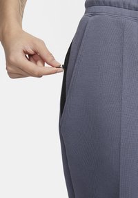 Grijze sportieve broek met een gestructureerde afwerking en een ritszak. Een hand reikt naar de rits van de zak, waardoor een contrasterende zwarte voering zichtbaar wordt.