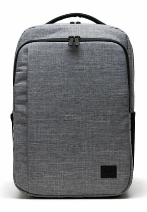 Sac à dos rectangulaire en tissu gris avec une poignée supérieure, une poche zippée à l'avant et un patch logo noir en bas à droite.