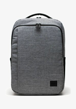 Sac à dos rectangulaire en tissu gris avec une poignée supérieure, une poche zippée à l'avant et un patch logo noir en bas à droite.