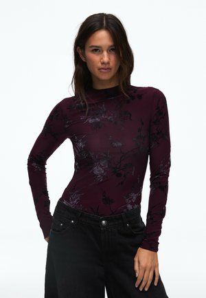 Femme aux cheveux foncés portant un haut violet ajusté à manches longues avec des motifs floraux noirs et un pantalon noir ample, debout avec une main sur la hanche.
