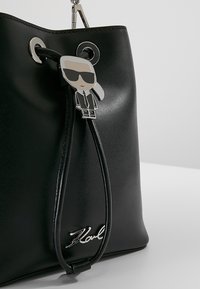 Sort læder bucket taske med sølvlogo, justerbar snørelukning og en vedhæng, der forestiller en figur med solbriller.
