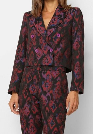 Vrouw draagt een bijpassend cropped blazer en broek met rood, paars en blauw abstract patroon en paarse knopen.