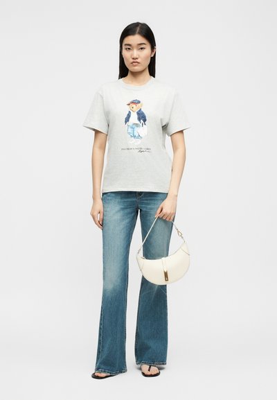 Γυναίκα που φοράει ανοιχτό γκρι μπλουζάκι με γραφικό αρκούδα, μπλε τζιν καμπάνα, μαύρα σανδάλια και κρατάει μια λευκή τσάντα σε σχήμα ημισελήνου.