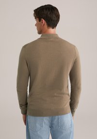Pulover lung cu mânecă lungă, tricotat, de culoare maro, cu guler și mansete cu nervuri, având o textură fină și un design ajustat. Purtați cu blugi de denim de culoare albastru deschis.
