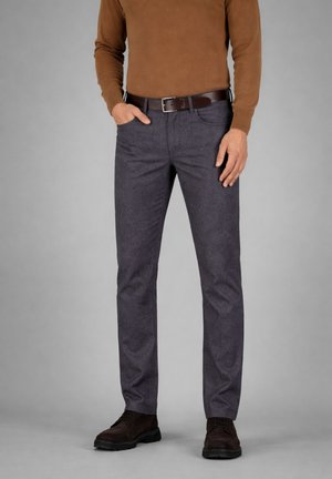 Uomo che indossa pantaloni grigi a gamba dritta, cintura marrone, maglione marrone e scarpe stringate marrone scuro su uno sfondo grigio semplice.