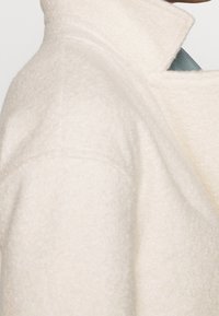 Cappotto in misto lana color crema, con colletto strutturato, tessuto testurizzato e una vestibilità sartoriale con dettagli di cuciture visibili sulla spalla.