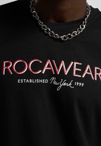 Fekete pamut póló, amelynek feltűnő piros és fehér "ROCAWEAR" logója van, finomabb szöveggel: "ESTABLISHED New York 1999", amelyet egy ezüst láncnyaklánc egészít ki.
