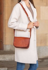 Borsa a tracolla in pelle marrone con texture liscia, forma rettangolare e tracolla regolabile. Indossata con un cappotto beige e jeans.