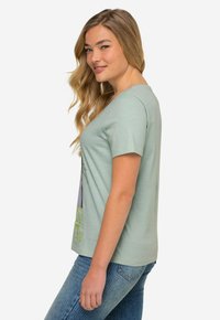 Camiseta verde menta de mangas cortas, corte relajado y cuello redondo. Presenta un estampado gráfico en la parte delantera. Combinada con vaqueros azules.
