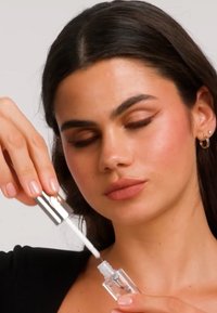 Kvinne med mørkt hår holder og fjerner applikatorstav fra et klart leppeglanstube mot en nøytral bakgrunn.