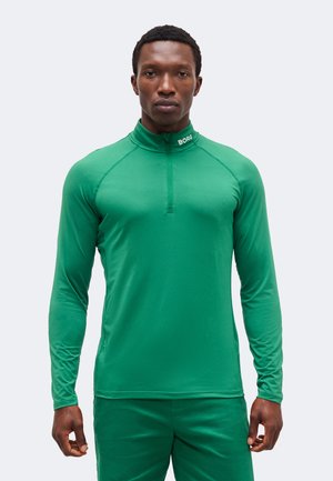 Άνδρας που φοράει πράσινο αθλητικό μακρυμάνικο μπλουζάκι με φερμουάρ στο ένα τέταρτο και τη λέξη "BORG" στο γιακά, στέκεται μπροστά σε απλό φόντο.