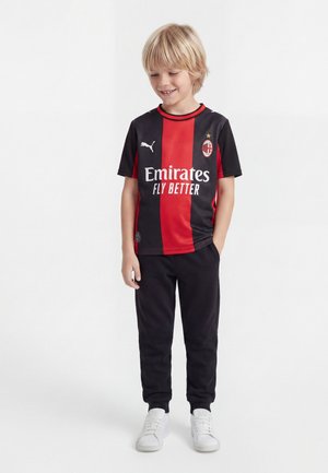 Băiat blond purtând tricou de fotbal AC Milan negru cu dungi roșii, pantaloni negri și adidași albi, stând cu mâinile în buzunare.