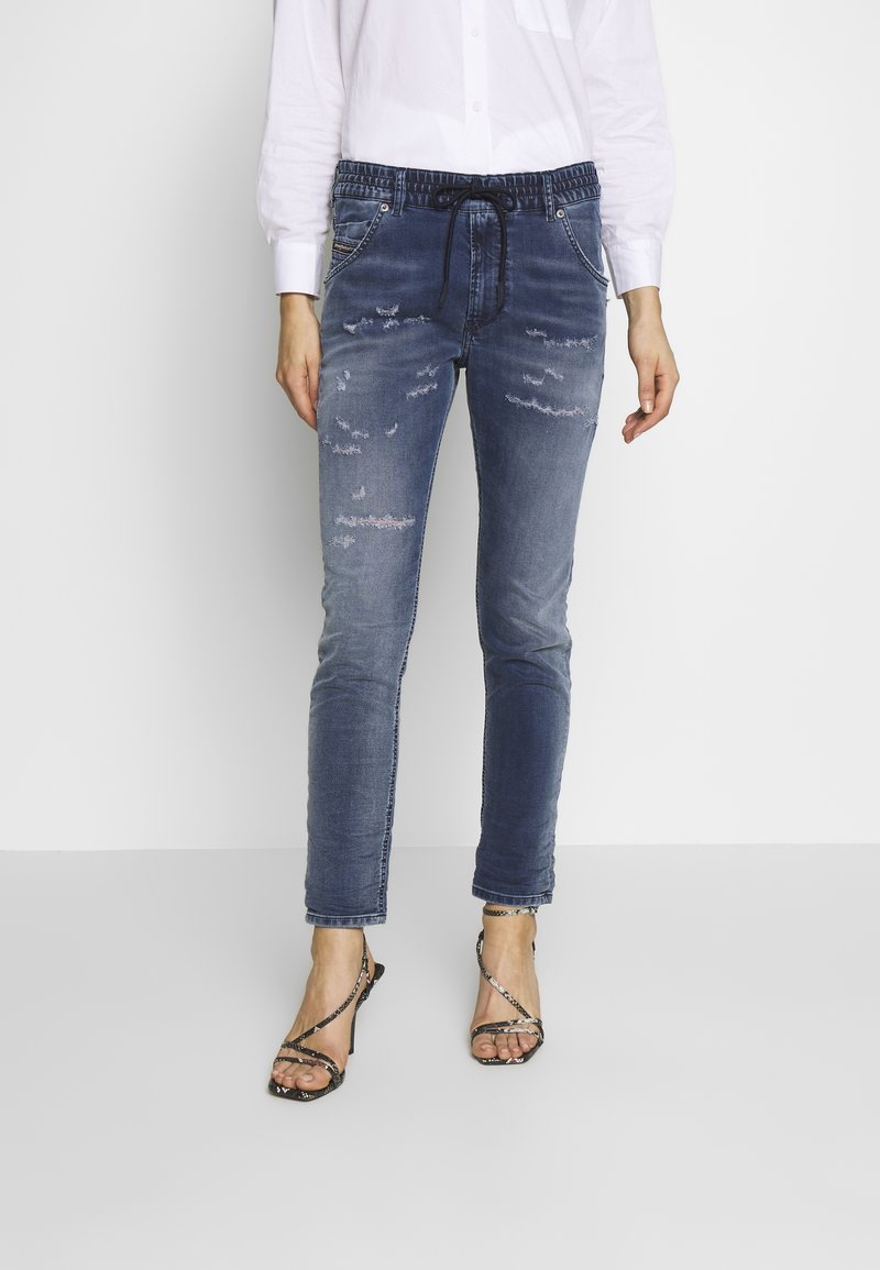 Nainen, joka on pukeutunut haalistuneisiin sinisiin kuluneisiin jogger-farkkuihin, joissa on naru vyötäröllä, ja mustiin remmiin korkokenkiin, seisoo yksinkertaisella taustalla.