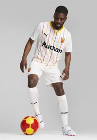 Uniforme de fútbol blanco con rayas verticales rojas y amarillas, con cuello en V y logotipo de patrocinador negro; el jugador está de pie sobre una pelota roja y amarilla.