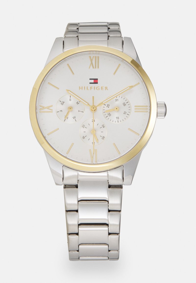 Reloj de acero inoxidable plateado con esfera blanca, acentos dorados, marcadores en números romanos y tres subesferas; pulsera de eslabones metálicos.
