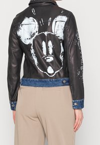 Giacca di pelle nera con dettagli in denim, caratterizzata da un grande design di un volto cartoonesco bianco sul retro. Chiusura con bottoni a pressione e dettagli ai polsini.