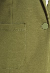 Tessuto verde oliva con una texture liscia, dotato di una tasca laterale e un pulsante rotondo decorativo. Dettagli di cucitura visibili lungo il bordo della tasca.