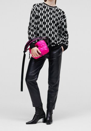 Person iført sort og hvid mønstret sweater, sorte læderbukser og sorte støvler med hæl, der holder en lille lyserød quiltet taske.