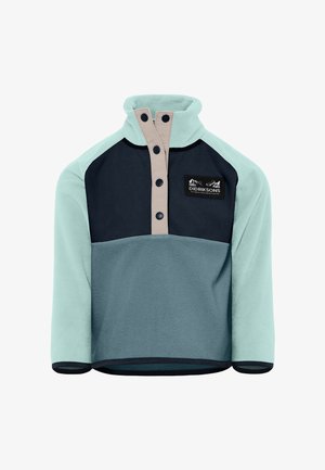 Jeune garçon debout, portant une veste en polaire à blocs de couleurs, un pantalon foncé et des baskets blanches, sur un fond clair uni.