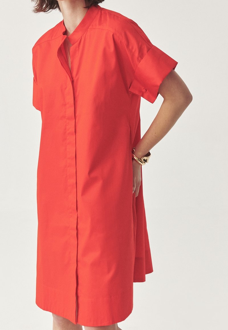 Femme portant une robe chemise rouge vif mi-longue aux genoux, à manches courtes et avec une patte de boutonnage cachée à l'avant, debout avec une main dans le dos.